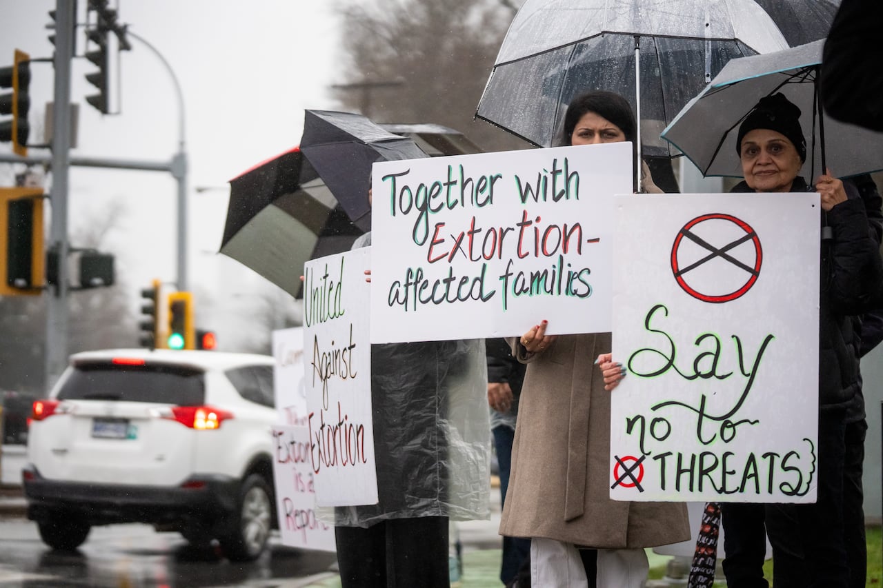 People hold signs and umbrellas on a rainy day. The signs have messages against extortion and threats.