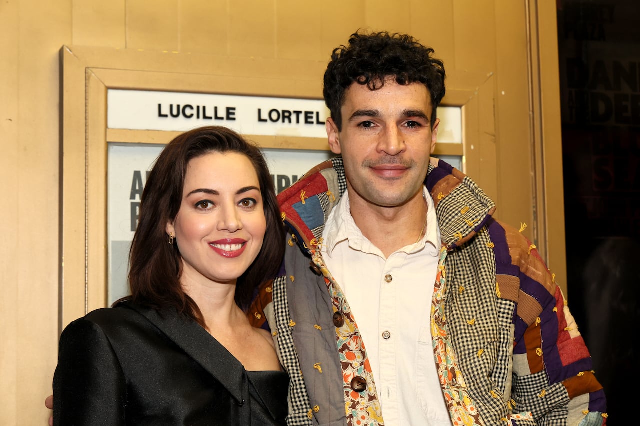 Woman in black sily jacket smiles alongside man in colourful patterned jacket in front of light wood wall. 