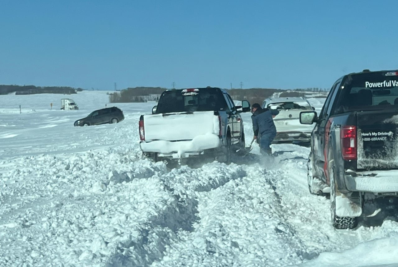 Vehicles are seen in the ditch on the side of the road. A man is trying to shovel out a truck/