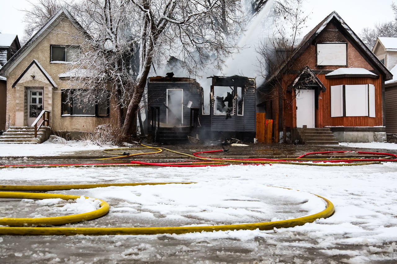 A burned house stands between two other homes. A hose pours water down onto the burned one.