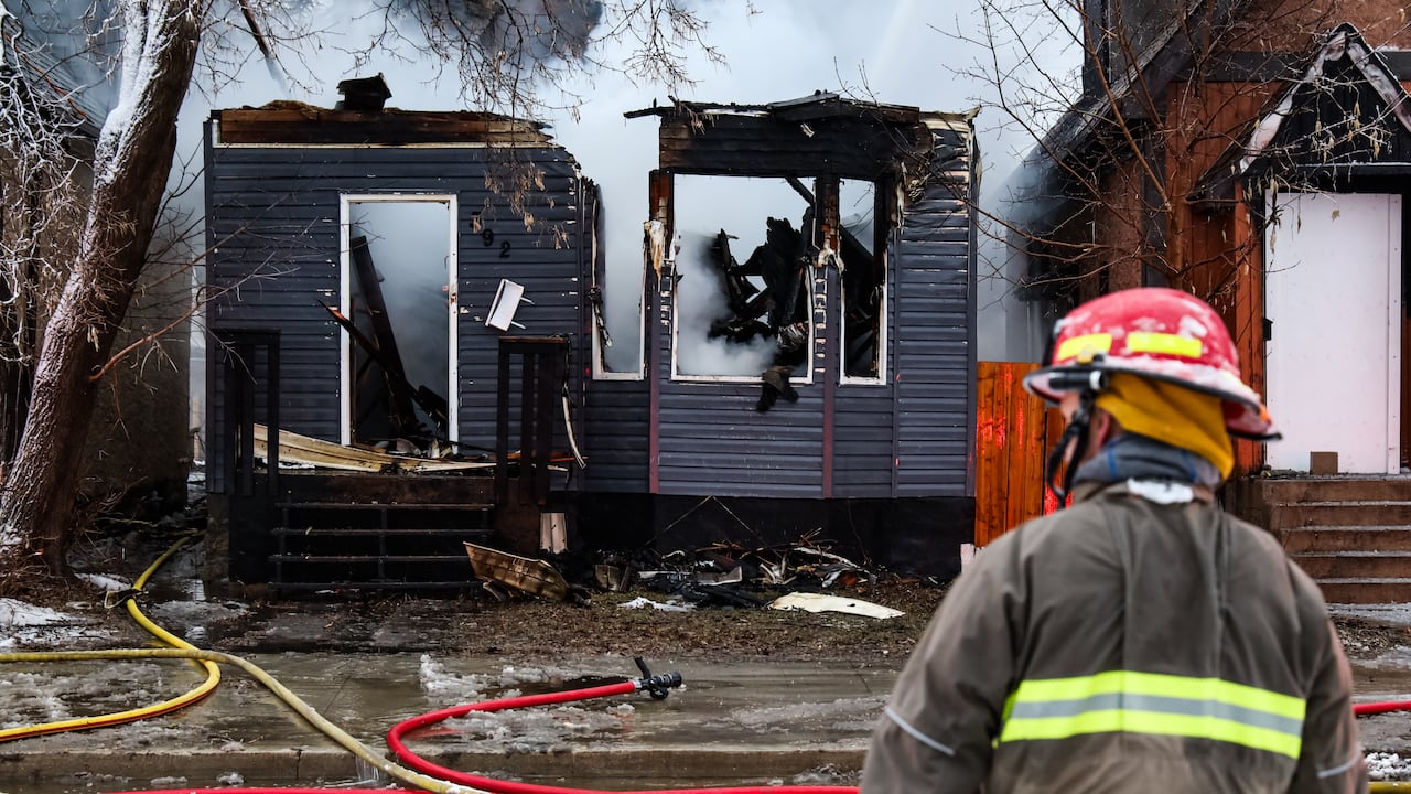 A firefighter stands in the foreground. A burned house is filled with smoke as water is sprayed from above.
