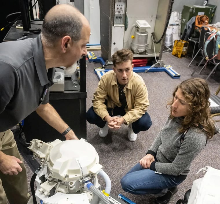 A man leans over a white tank with hoses connected to it while two others squat nearby, listening. 