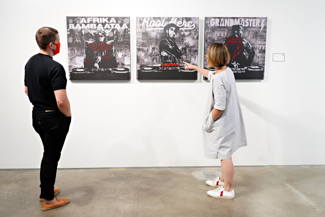 A man and a woman stand in a gallery in front of three separate portraits on a wall.