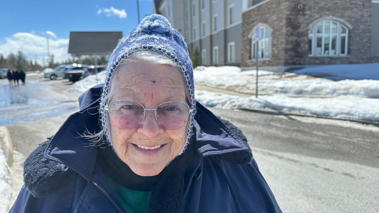 An elderly woman wearing a blue winter coat and blue fleece hat with white snowflakes in front of a recently built nursing home