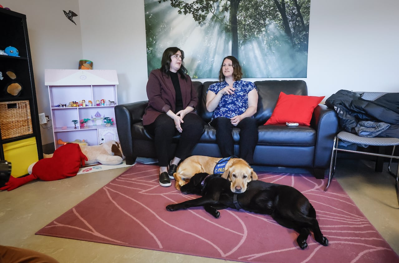 Two women sit on a couch, with two dogs lounging on the carpet at their feet.