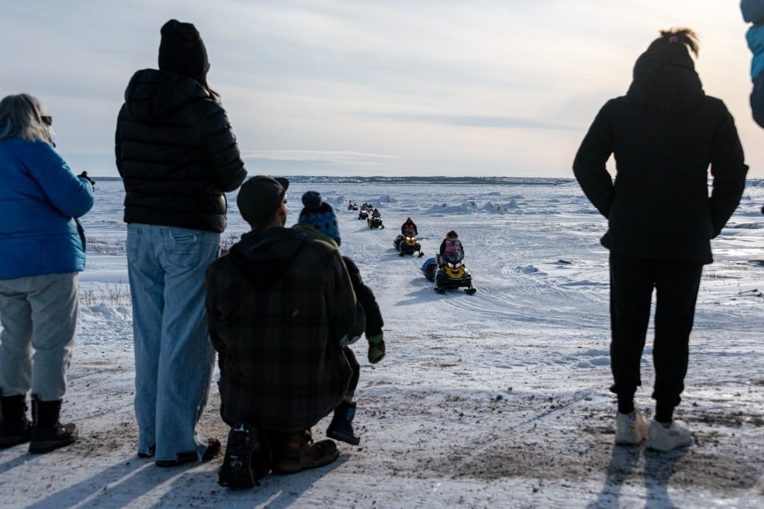 A group of people, including a child, looking toward a column of people driving snowmobiles on a snowy landscape.