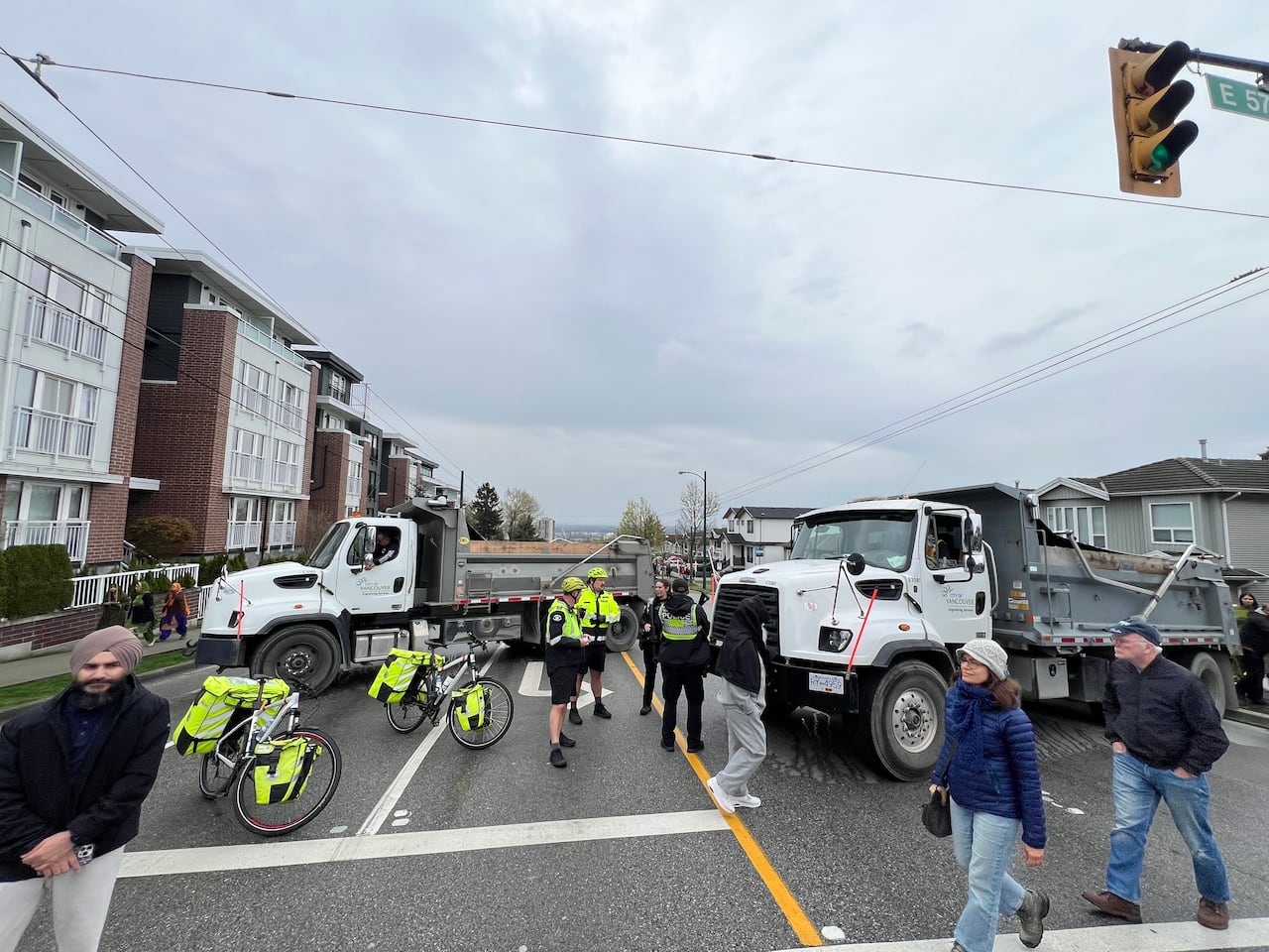 Two dump trucks block a residential intersection as pedestrians and police officers stand nearby.