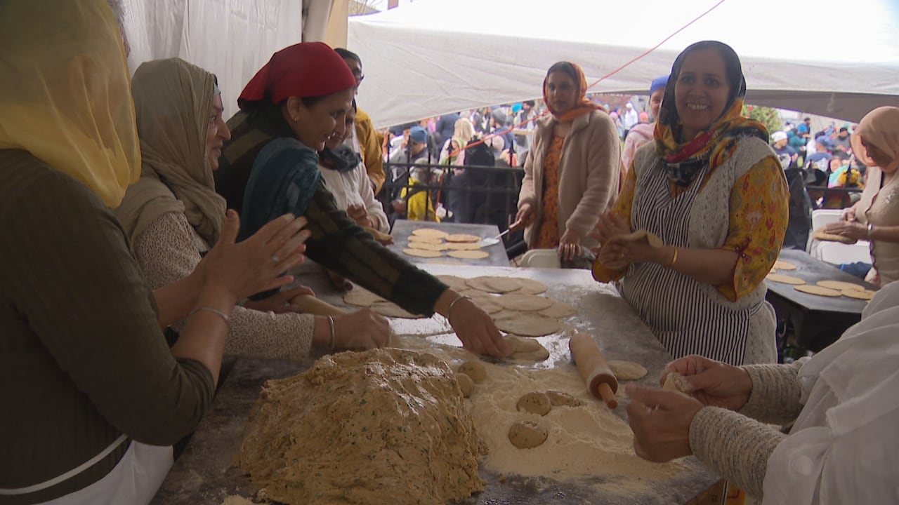A group of women roll and prepare dough together at a table inside a tent.