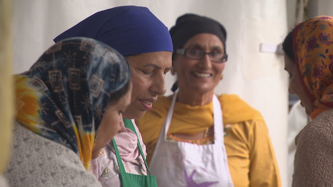A group of women wearing headscarves and aprons prepare food together, smiling and working closely.