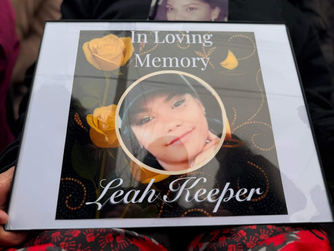 A framed picture showing the face of a woman surrounded by flowers. Overlaid text reads 'In Loving Memory' and 'Leah Keeper.'