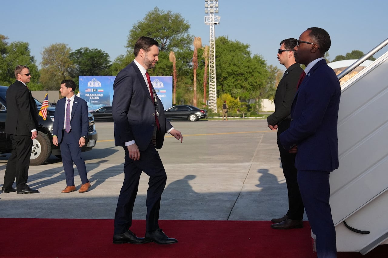 A man in a suit walks on a red carpet towards stairs to an airplane.