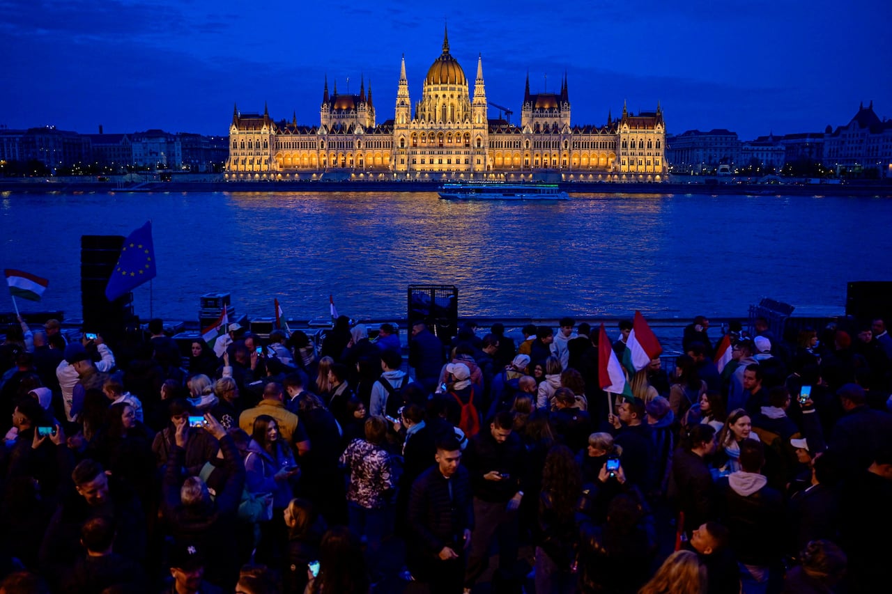Demonstrators gather near the shore of a river as a building is seen in the background.