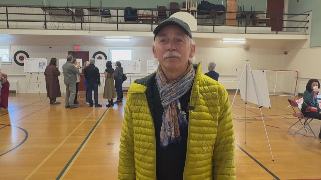 A man wearing ac cap, blue scarf and yellow puffer jacket stands in front of the camera.