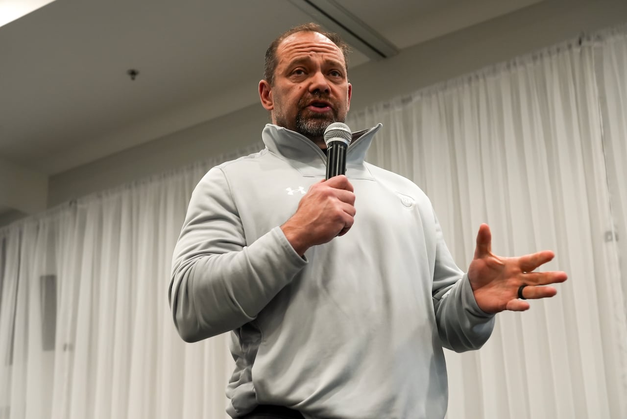 A man in a grey, half-zip athletic sweater stands on a stage with a microphone. 