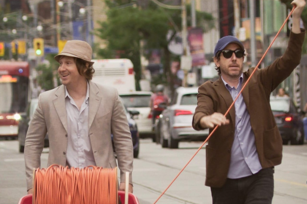 Two men, one in a beige hat and suit jacket and shirt and one in sunglasses a blue ball cape, brown jacket and light purple shirt walk down the street with a coil of orange cord in a red wheel barrel. It's a seen from the movie: Nirvanna the Band the Show the Movie.