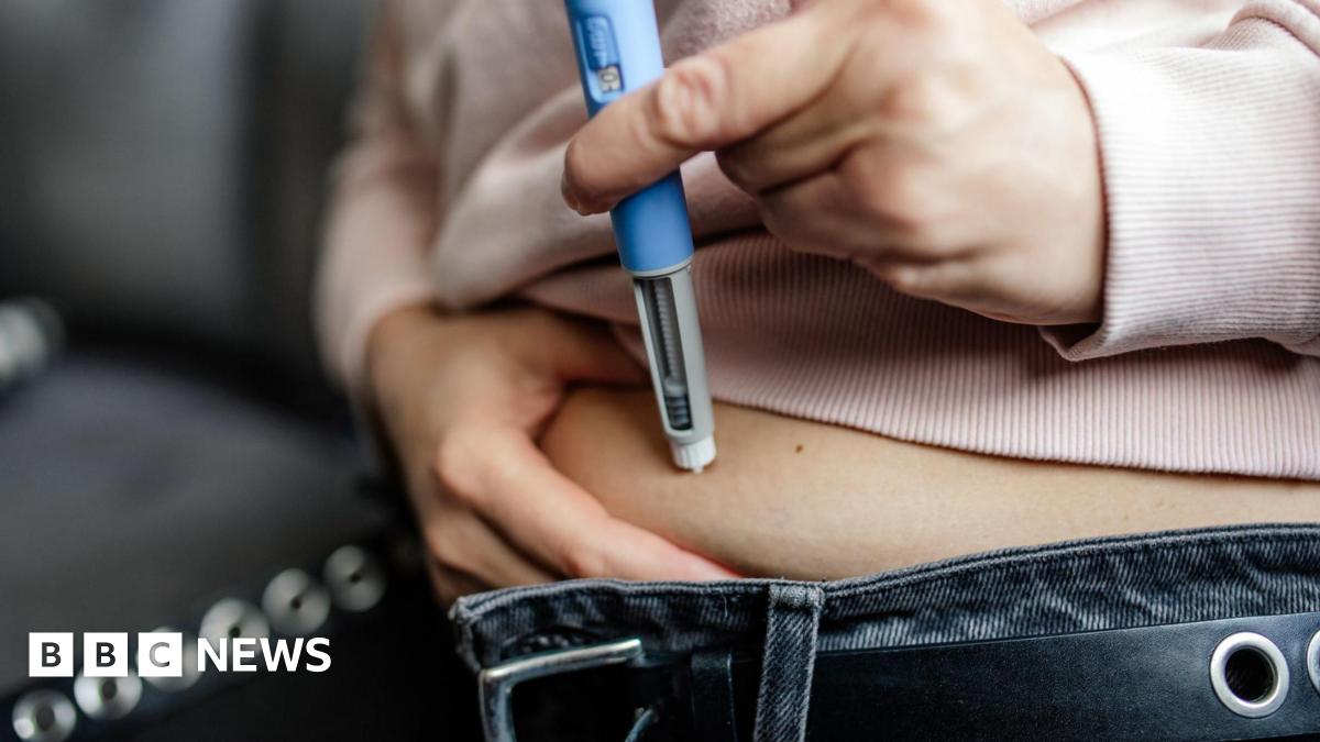 A woman, whose face and upper torso is not shown, injects a weight-loss drug via an injection pen into her abdomen, with the belt of her blue jeans unbuckled and her pink jumper lifted up slightly