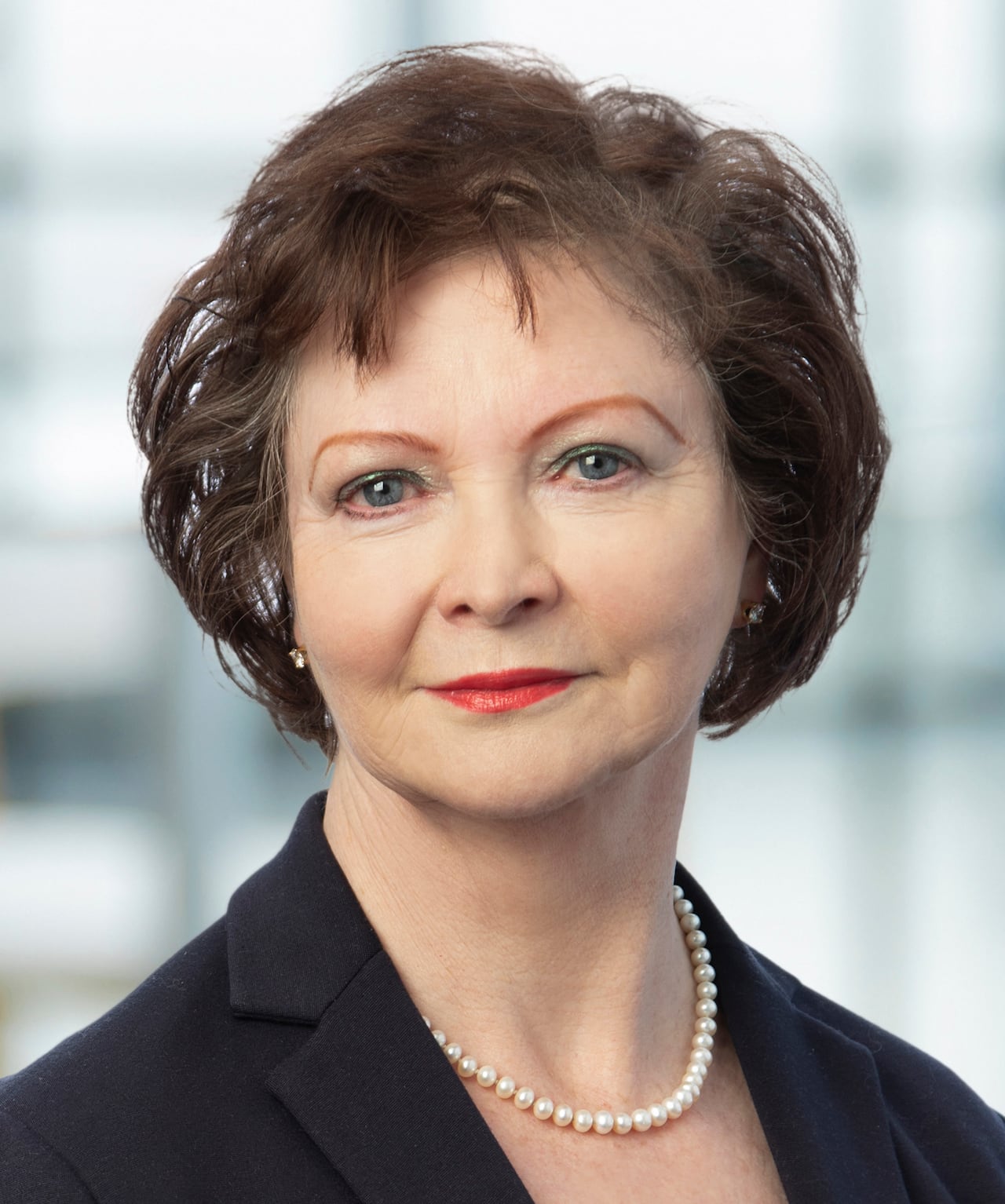 A woman with brown hair, wearing a dark blazer and a pearl necklace poses for a headshot