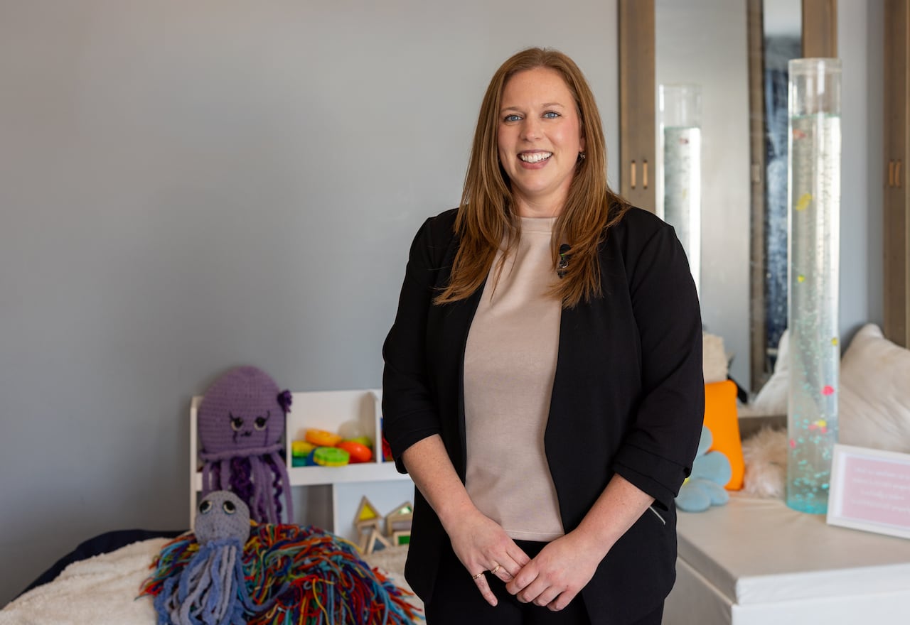 A smiling woman with long brown hair stands in a playroom featuring colorful toys