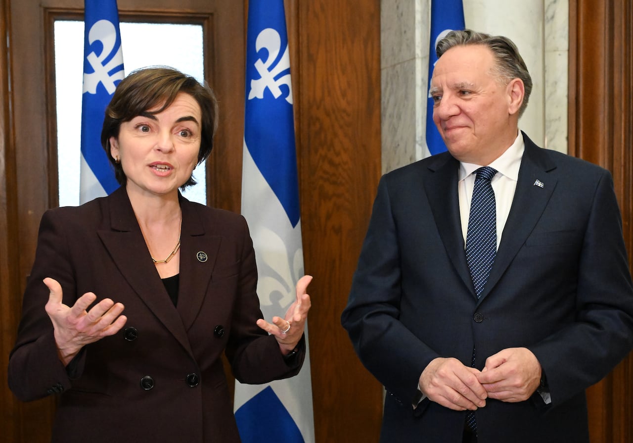 A woman with short hair and wearing a maroon blazer stands next to a man wearing a navy blazer. He's smiling at the woman.