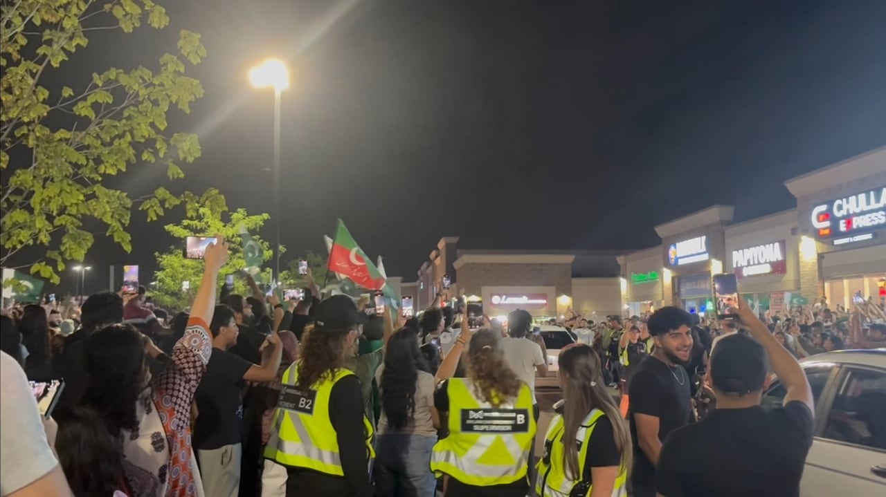 A large crowd of people outside of a row of shops, three people in the front wear high visibility vests.