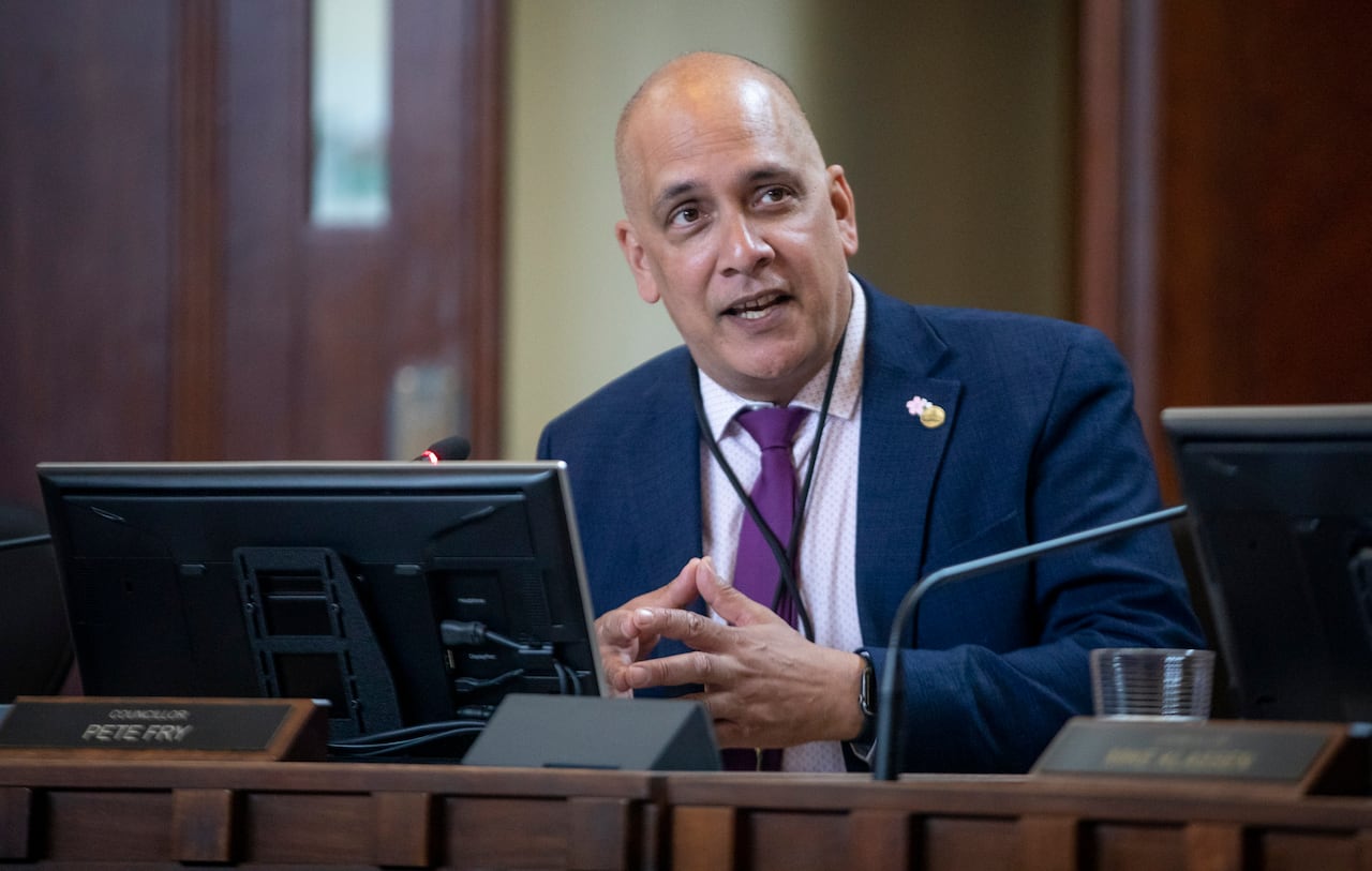 A man wearing a purple tie and blue suit jacket speaks in council chambers.