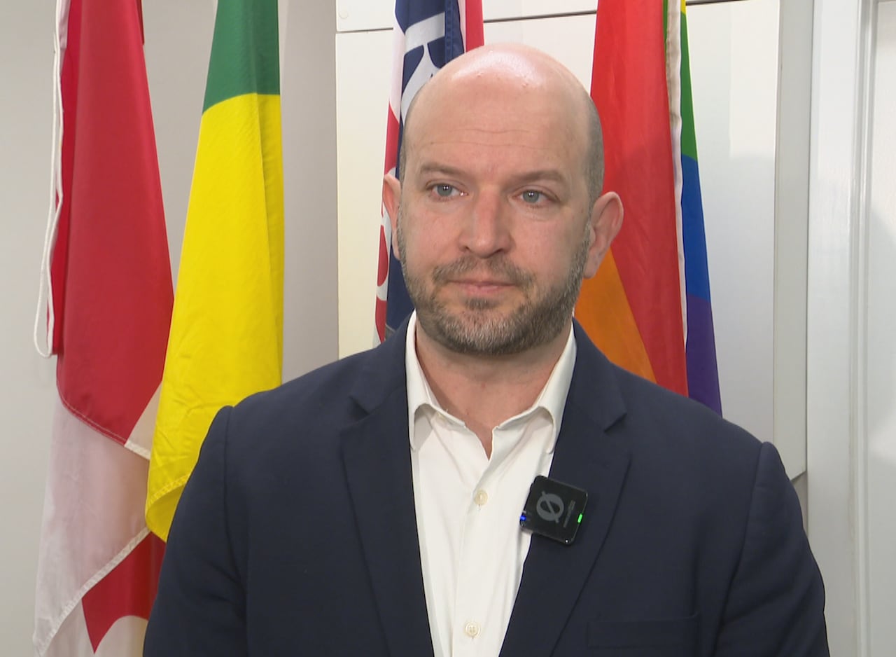 A man wearing a dark blazer stands in front of flags.