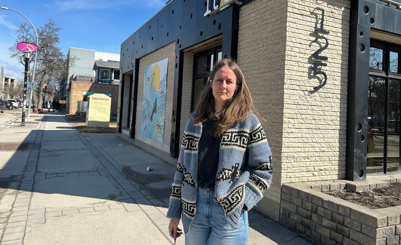 woman with long brown hair wears a blue, black and white knit sweater and stands on a city street outside a building that says "RAY" on its sign