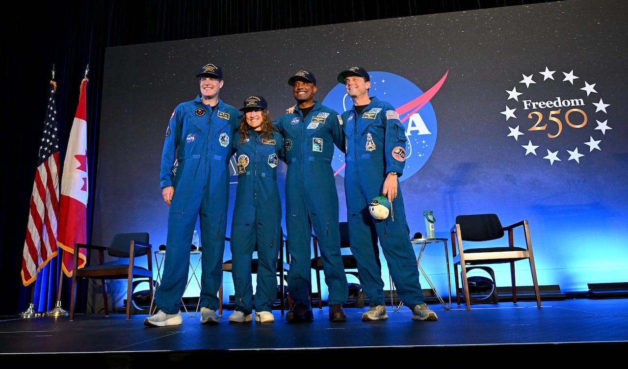 Four people in blue jumpsuits and hats wave from a stage.
