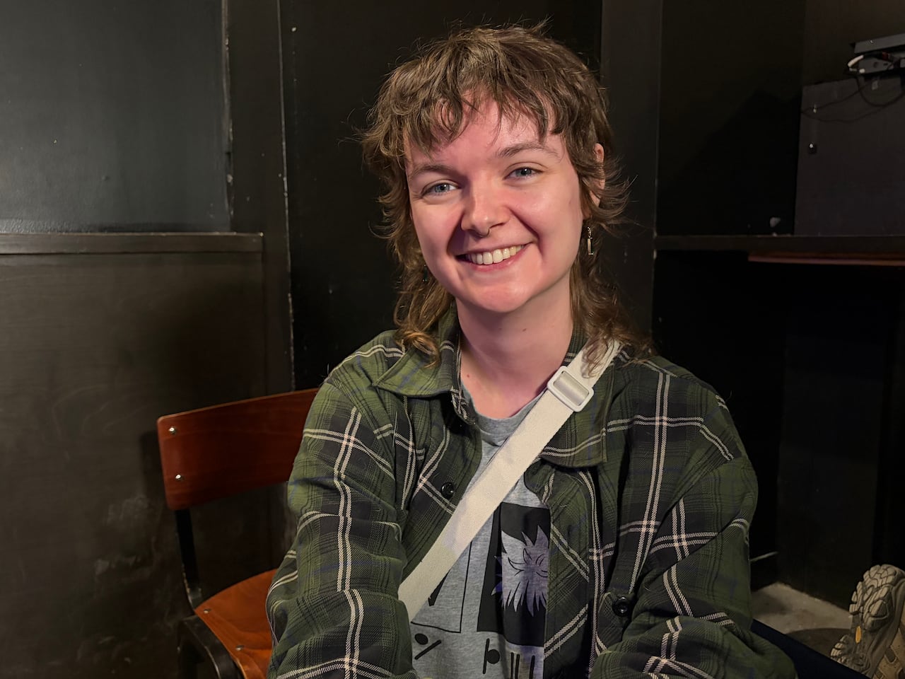 A person in a green plaid shirt and grey t-shirt is seated at a chair.