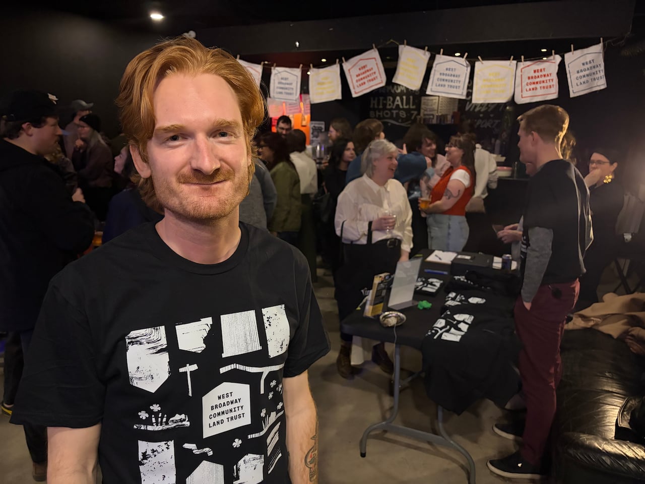 A man in a black t-shirt with white drawings stands in the foreground while a bunch of people attending the party mingle in the background.