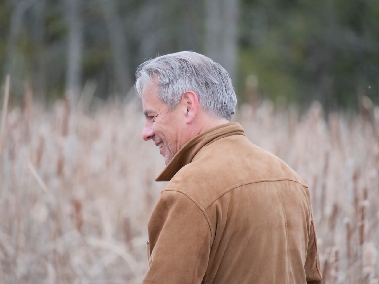 Drew Hayden Taylor in three quarter profile in a beige taupe jacket, standing in a wheat field.