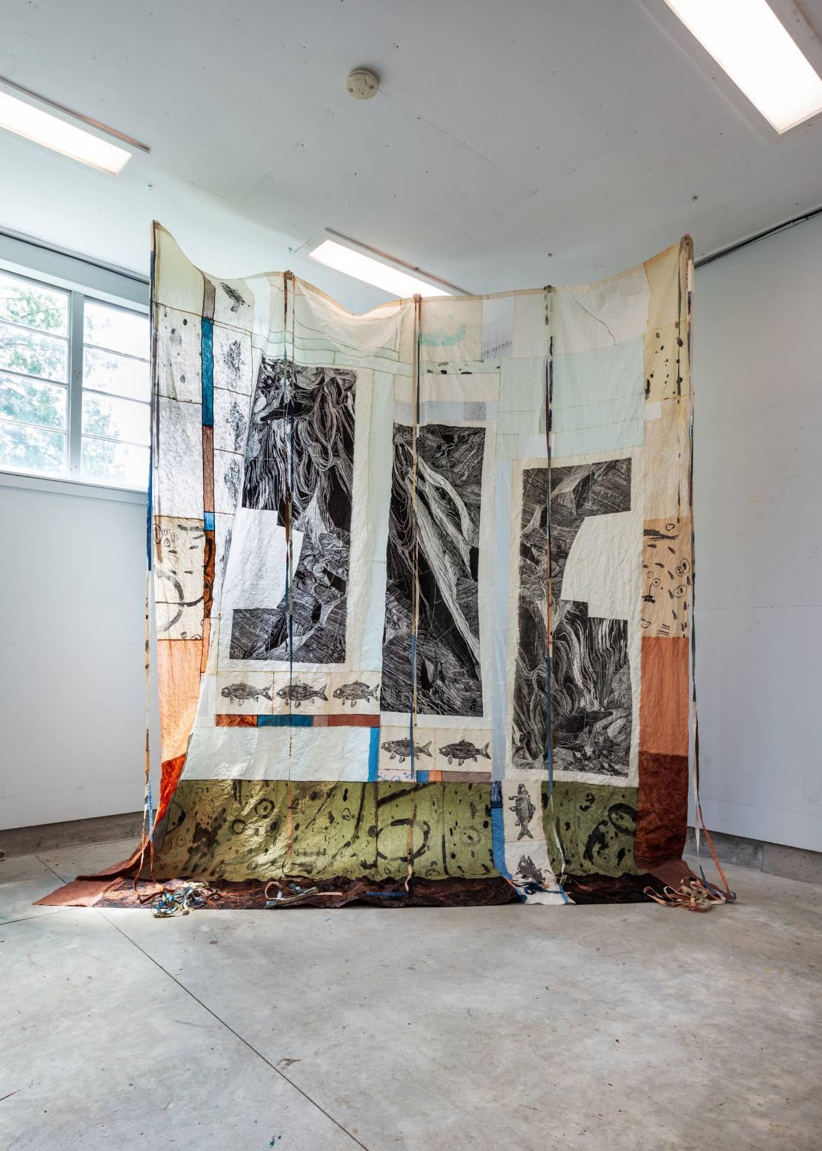 Textile artwork installed in empty white walled room. It's nearly as tall as the ceiling and is made of a patchwork of fabrics and designs.