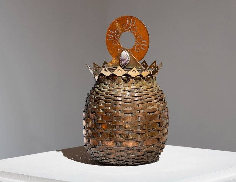 Sculptural artwork photographed in blank grey-walled room. Resembles a woven basket topped with a pointy crown of metal embellished with an inset shell.