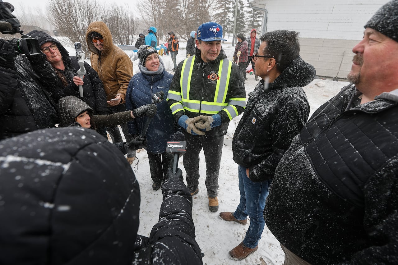 A man wearing a high-visibility best and gloves standing in front of several people holding microphones, He's speaking with another man wearing winter gear.