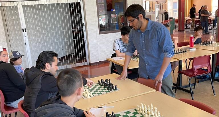 A man in glasses and a blue shirt stands by a table across from other people with a chess board between them.