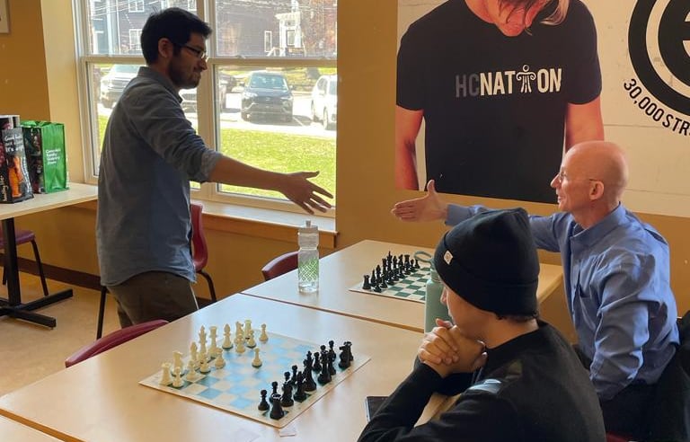 A man in glasses and a blue shirt extends his hand to a man in a blue shirt with a table and a chess board between them.