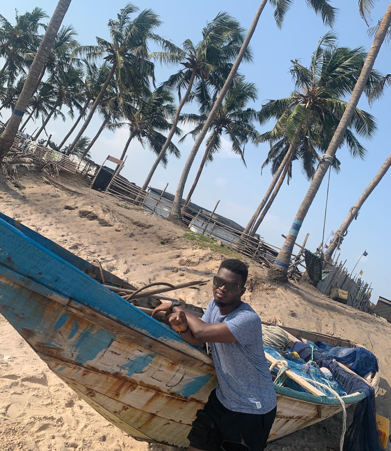 A smiling man leans against a boat on a sandy beach.
