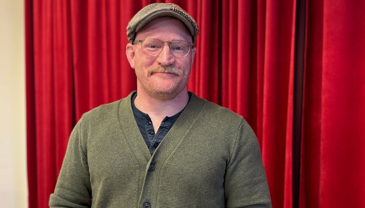 A man with a blonde moustache with wears a green cardigan and flat cap, with red curtains behind him