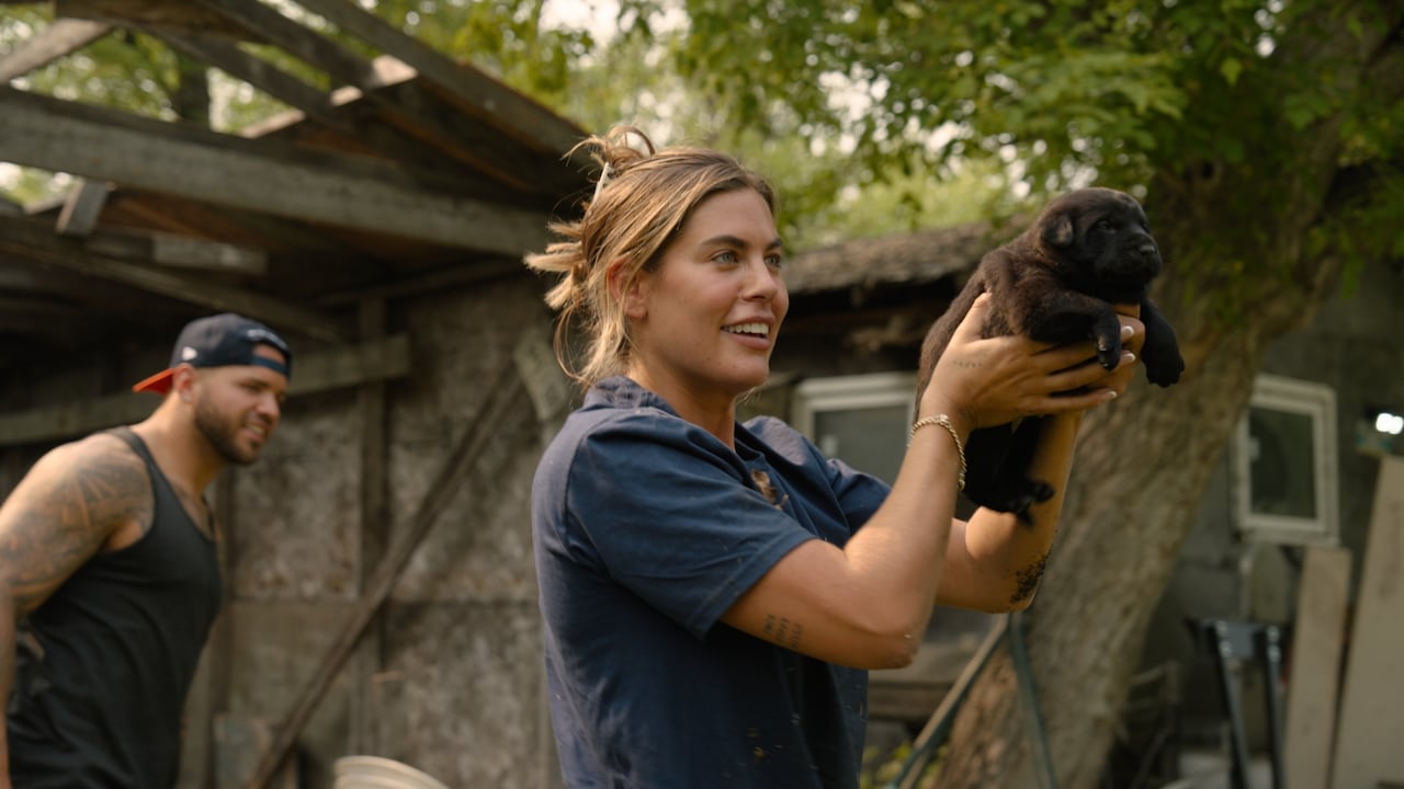 A man and a woman in front of a home. The woman holds up a small, black puppy.