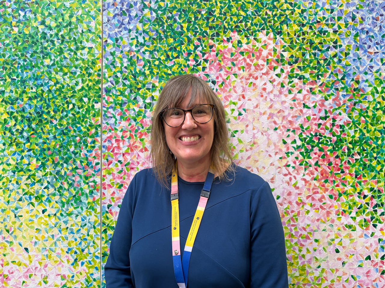A woman wearing glasses and a lanyard stands smiling in front of a colourful painted wall.