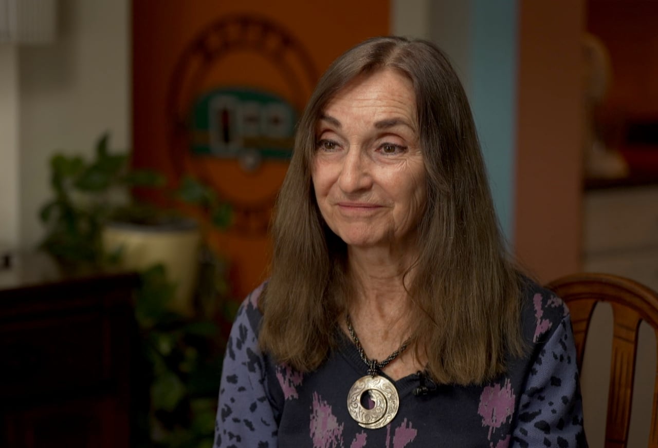 Joan Prowse is sitting in her living room wearing a purple top and necklace.