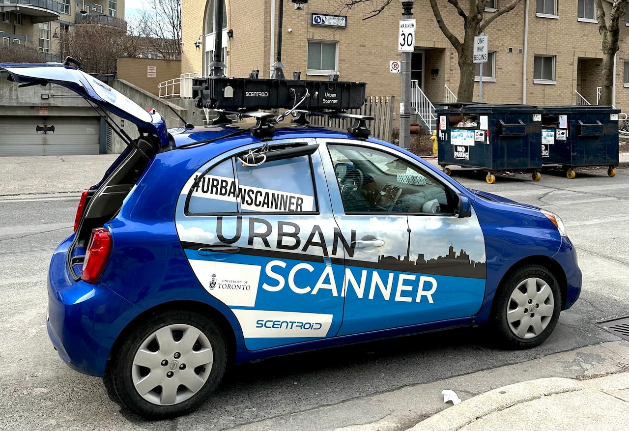 Small blue car with "Urban Scanner" printed on its side is parked in a residential street