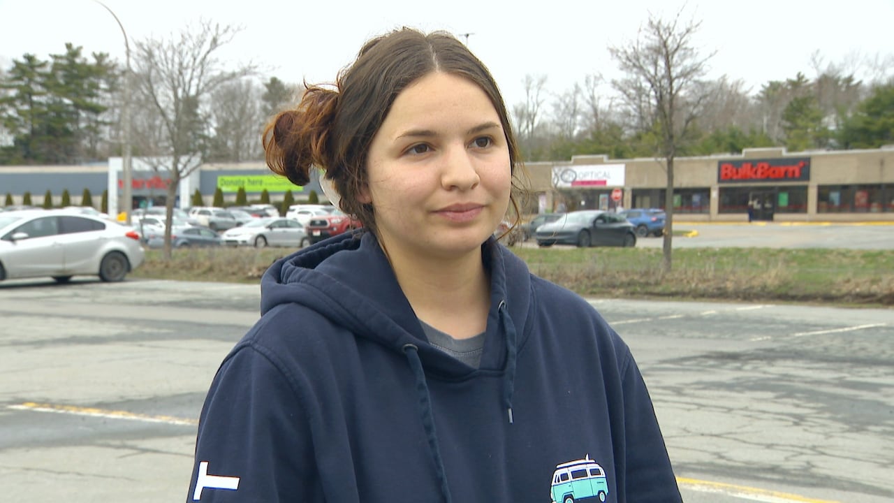 A young white woman with brown hair pulled back in a bun wears a navy hooded sweatshirt. She is standing outside, with a strip mall behind her