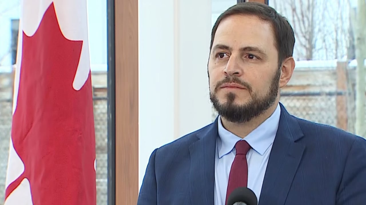  A man in a blue suit stands in front of a window near a Canadian flag.