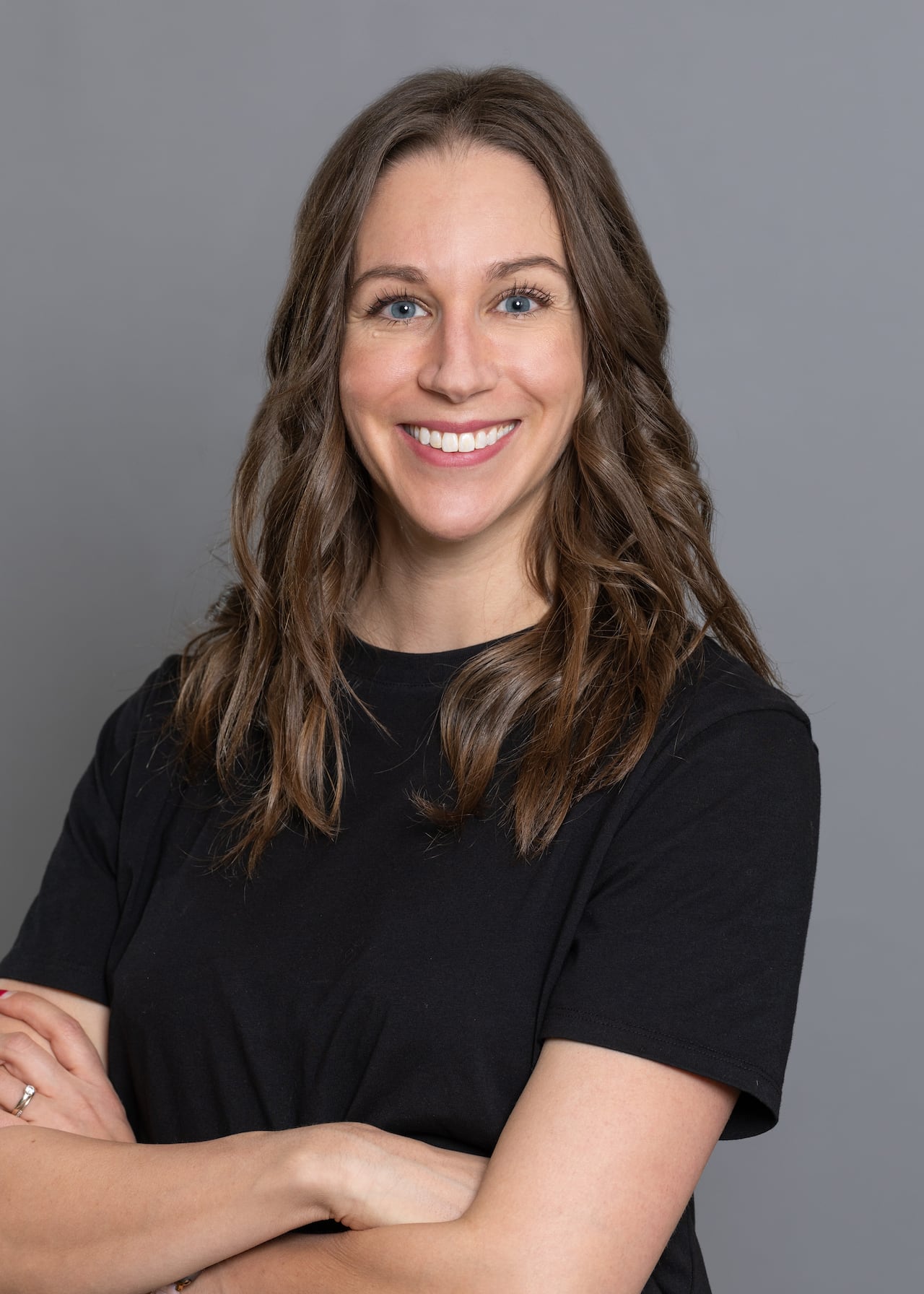 A woman with wavy brown hair has her arms crossed and is wearing a black short-sleeved shirt as she smiles at the camera