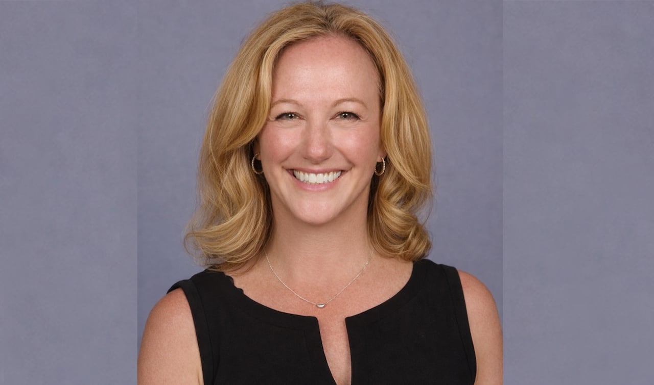 A portait of a smiling woman in a dark, sleeveless top and standing against a blue-grey backdrop.