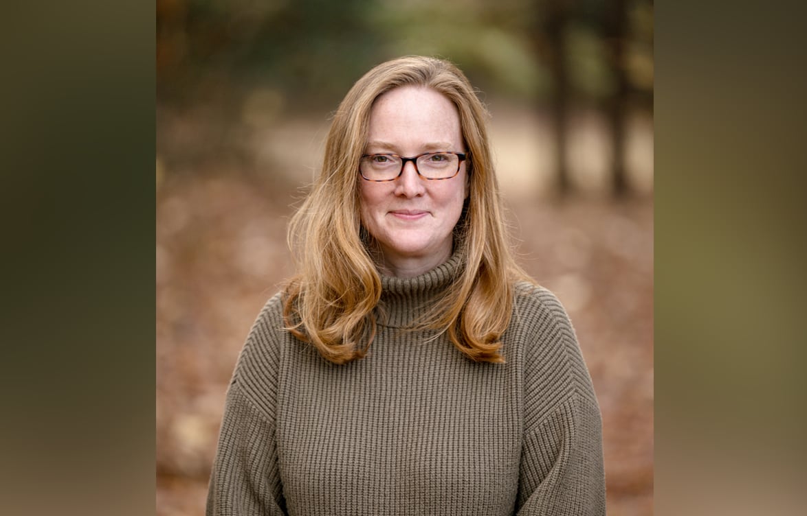 A portrait of a smiling woman with glasses and a light brown sweater standing outdoors, a forest setting out of focus behind her.