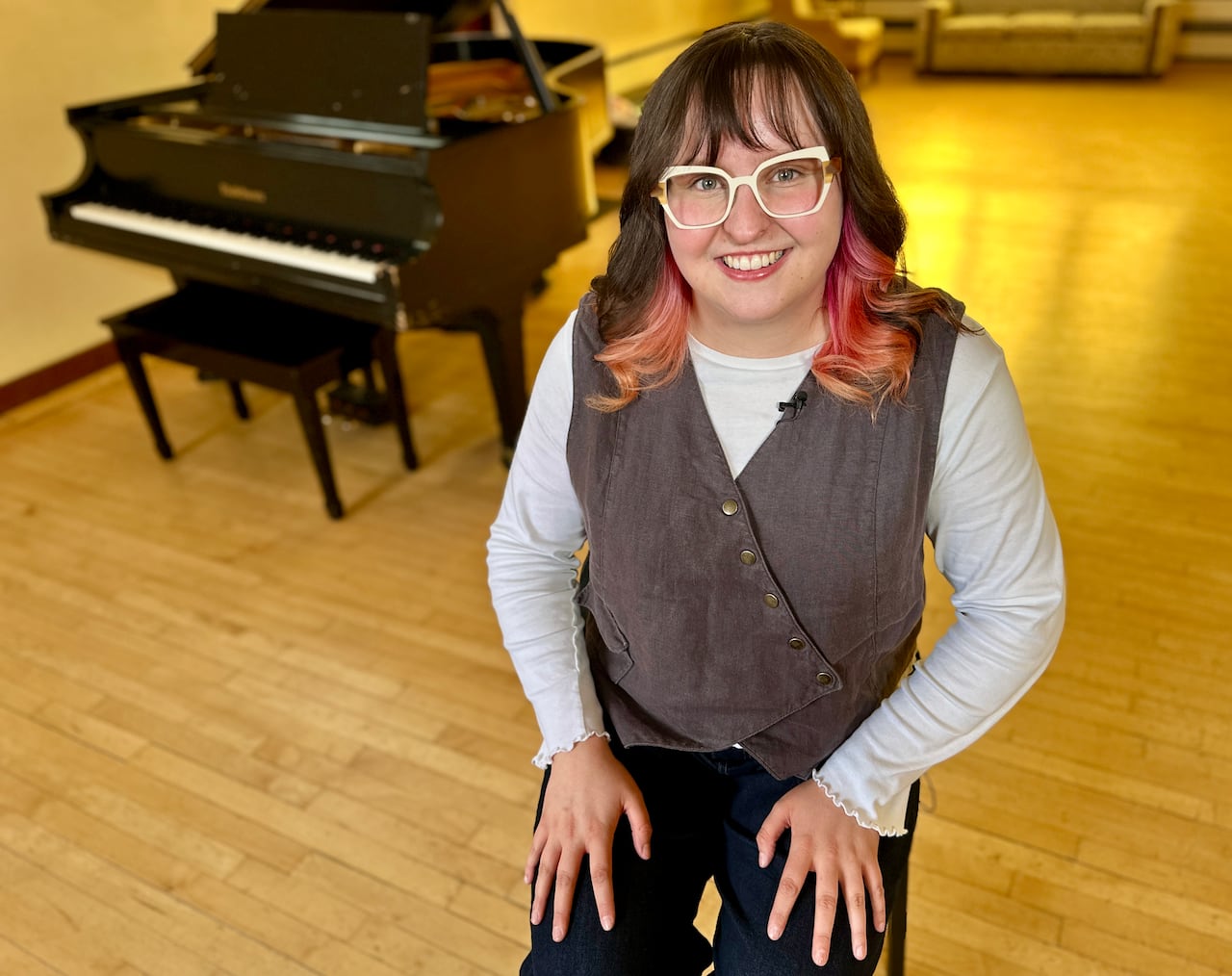 A young woman with white rimmed glasses and a big smile sits next to a baby grand piano.