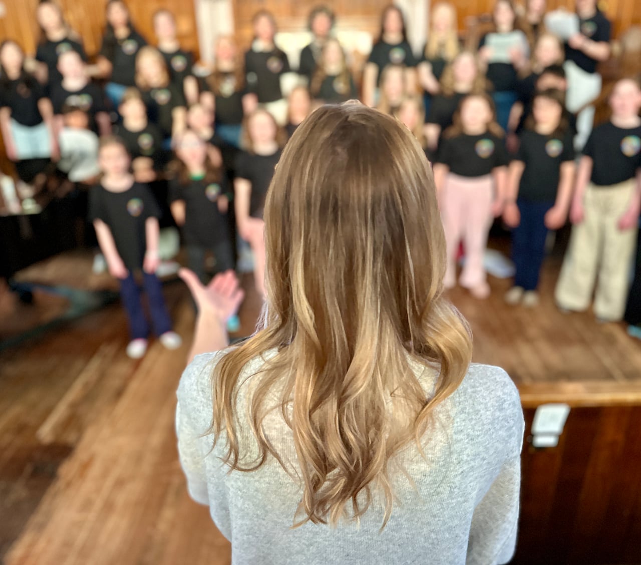 The back of a blonde head and hand looks out to direct a sea of singers on the steps in a church.