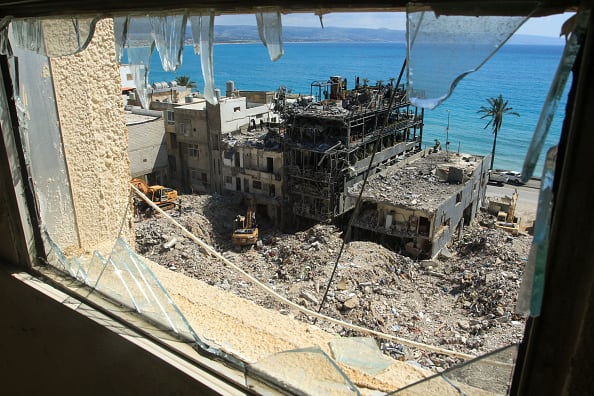 Diggers remove the rubble of buildings destroyed in airstrikes. In the background are the blue waters of an ocean.